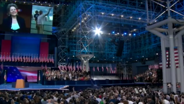 Eine große Menschenmenge vor einer Bühne mit einem Rednerpult, Mikrofon, Flaggen, einem Bildschirm und Beleuchtungsausrüstung, die auf eine Präsidenten-Debatte warten.