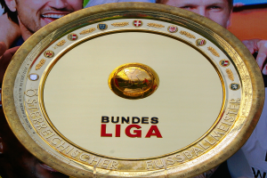 Ein Mann hält ein goldenes Schild mit der Aufschrift 'Bundesliga' hoch, im Hintergrund eine Fahne mit drei Personen.