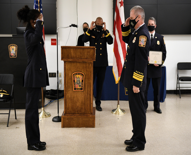 Gruppe uniformierter Personen in Masken vor einem Podium mit Mikrofon, umgeben von Fahnen, Stühlen, Tischen und einer Wand, mit einer Person, die ein Zertifikat während einer Amtseinführung für einen neuen Feuerchef hält.