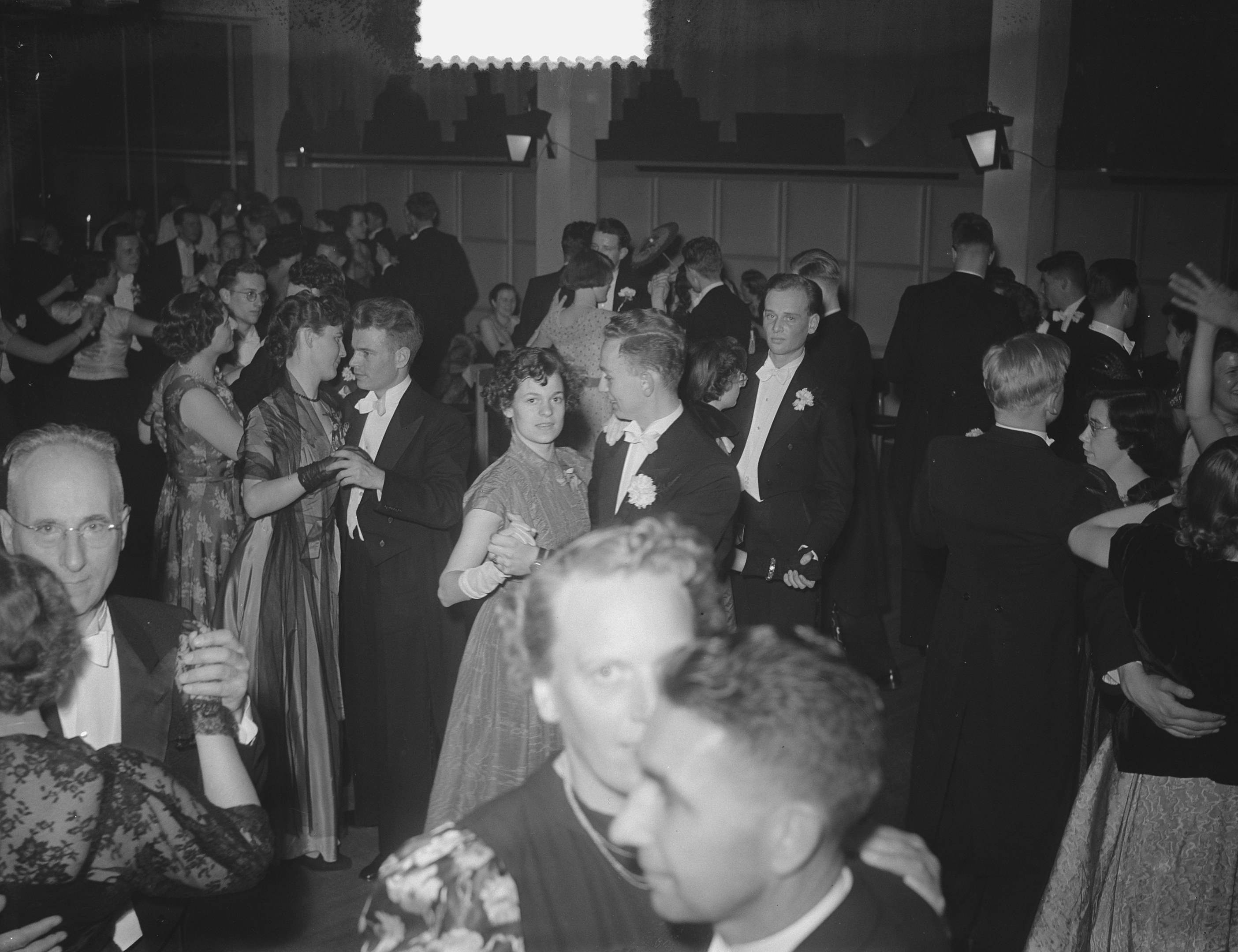 Eine Gruppe von Menschen verschiedenen Alters, die auf einer Party in einem Schwarz-Wei├č-Setting tanzen, einige halten Gl├Ąser, mit beleuchteter Wand im Hintergrund.