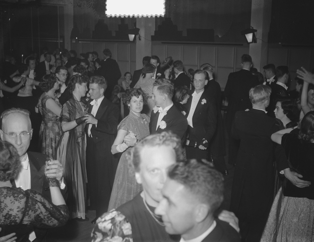 Eine Gruppe von Menschen verschiedenen Alters, die auf einer Party in einem Schwarz-Wei├č-Setting tanzen, einige halten Gl├Ąser, mit beleuchteter Wand im Hintergrund.