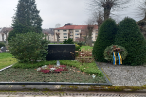 Ein Weltkriegsgedenkmal für Opfer steht in einem Park, geschmückt mit einem Kränzchen und umgeben von Grün, mit Gebäuden im Hintergrund.