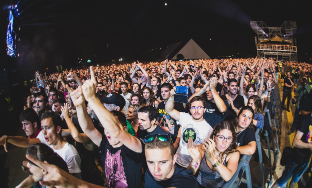 Menschenmenge auf einem Musikfestival mit erhobenen Händen, sitzend und stehend, Metallzaun auf der rechten Seite, Gebäude und Lichter im Hintergrund unter einem dunklen Himmel, mit einem Mann, einer Frau und anderen im Vordergrund.