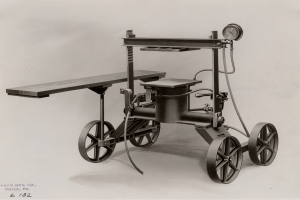 Ein altes Schwarz-Weiß-Foto einer Autoklavmaschine mit zylindrischem Körper, einem langen Arm und einem Druckmesser, die auf einem hölzernen Tisch mit vier Rädern stehen.