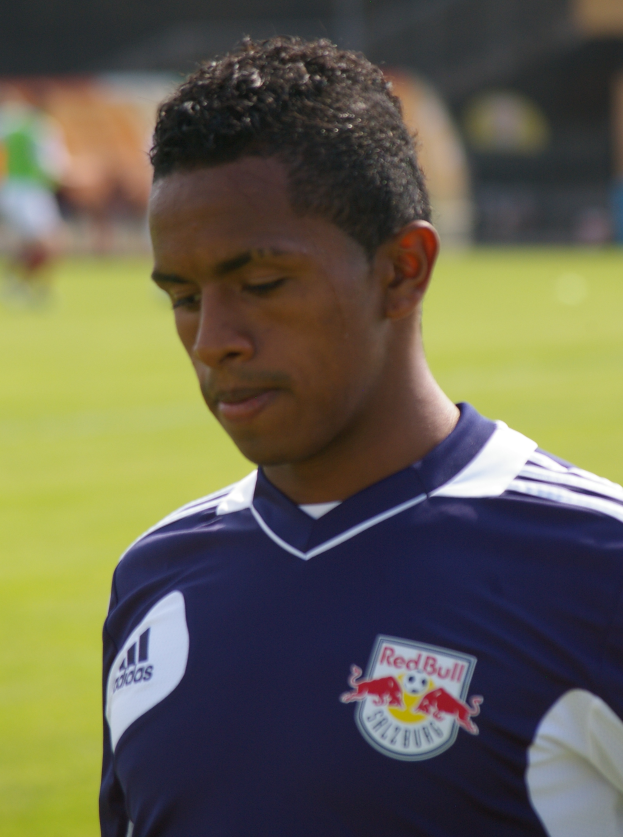 Ein junger Mann in einem blauen T-Shirt steht auf einem Fußballfeld, mit kurzen, dunklen Haaren und einem entschlossenen Gesichtsausdruck, der als Flügelspieler mit einem unscharfen Hintergrund erscheint.
