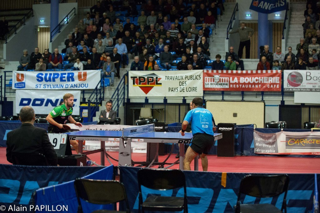 Indoor-Tischtennis-Match mit zwei Spielern und mehreren stehenden Zuschauern, sitzenden Publikum, leeren Stühlen und Werbetafeln in einer Stadionatmosphäre.