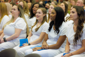 Eine Gruppe von Frauen in weißen Kleidern sitzt in einem Hörsaal, jede hält ein blaues Objekt, mit einem unscharfen Hintergrund, der die Frauen betont, die scheinbar Teil eines Pflegestudiums sind.