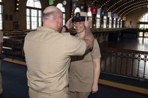 Ein Mann in Uniform heftet einer lachenden Frau eine Medaille an den Kopf, im Hintergrund sind Bänke, ein Geländer, Flaggen, Lichter, Fenster und eine Wand zu sehen.
