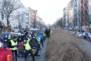 Eine große Gruppe von Menschen in Masken und Sicherheitswesten fährt Fahrräder eine straße mit Bäumen, Gebäuden mit Fenstern, Laternenmasten und Texttafeln entlang, während Fahrzeuge die Straße teilen und trockenes Gras die rechte Seite unter einem klaren blauen Himmel einnimmt.