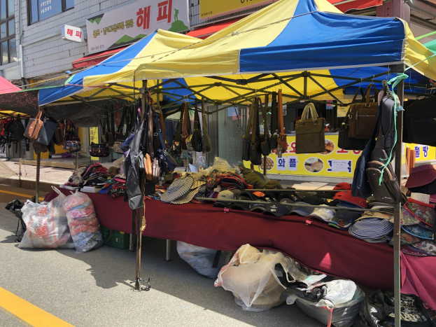 Ein Straßenmarkt mit Zelten, die Taschen, Hüte und andere Gegenstände ausstellen, mit Gebäuden und Texttafeln im Hintergrund.