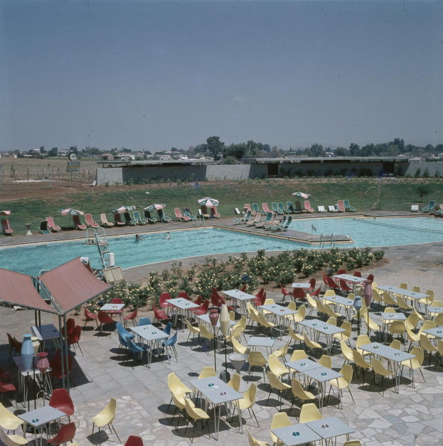 Ein Freibad mit Tischen, Stühlen und Sonnenschirmen, umgeben von Blumen, einem Zaun, einer Wand, Bäumen, Gebäuden und einem bewölkten Himmel.