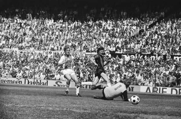 Schwarz-weiß-Foto von Männern, die auf einem Feld Fußball spielen, mit Zuschauern im Hintergrund und einer "Bundesliga 1978-79 Ajax 1-1 Hannover 96"-Fahne.