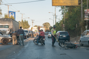 Gruppe von Menschen um ein verunglücktes Motorrad am Straßenrand mit mehreren Fahrzeugen, darunter ein Lastwagen, im Hintergrund und Bäumen, Pfählen, Laternen, Schildern und Himmel.