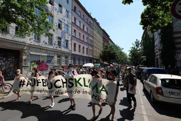 Eine Gruppe von Menschen hält ein Transparent und geht eine Straße in Berlin entlang, mit parkenden Autos auf der rechten Seite, einem Fahrradfahrer auf der linken Seite und Gebäuden, Bäumen und einem Schild im Hintergrund.