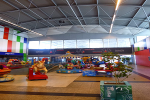 Amusement rides, a potted plant, scattered toys, informational boards, walls, ceiling lights, and metal poles in a large shopping mall interior.