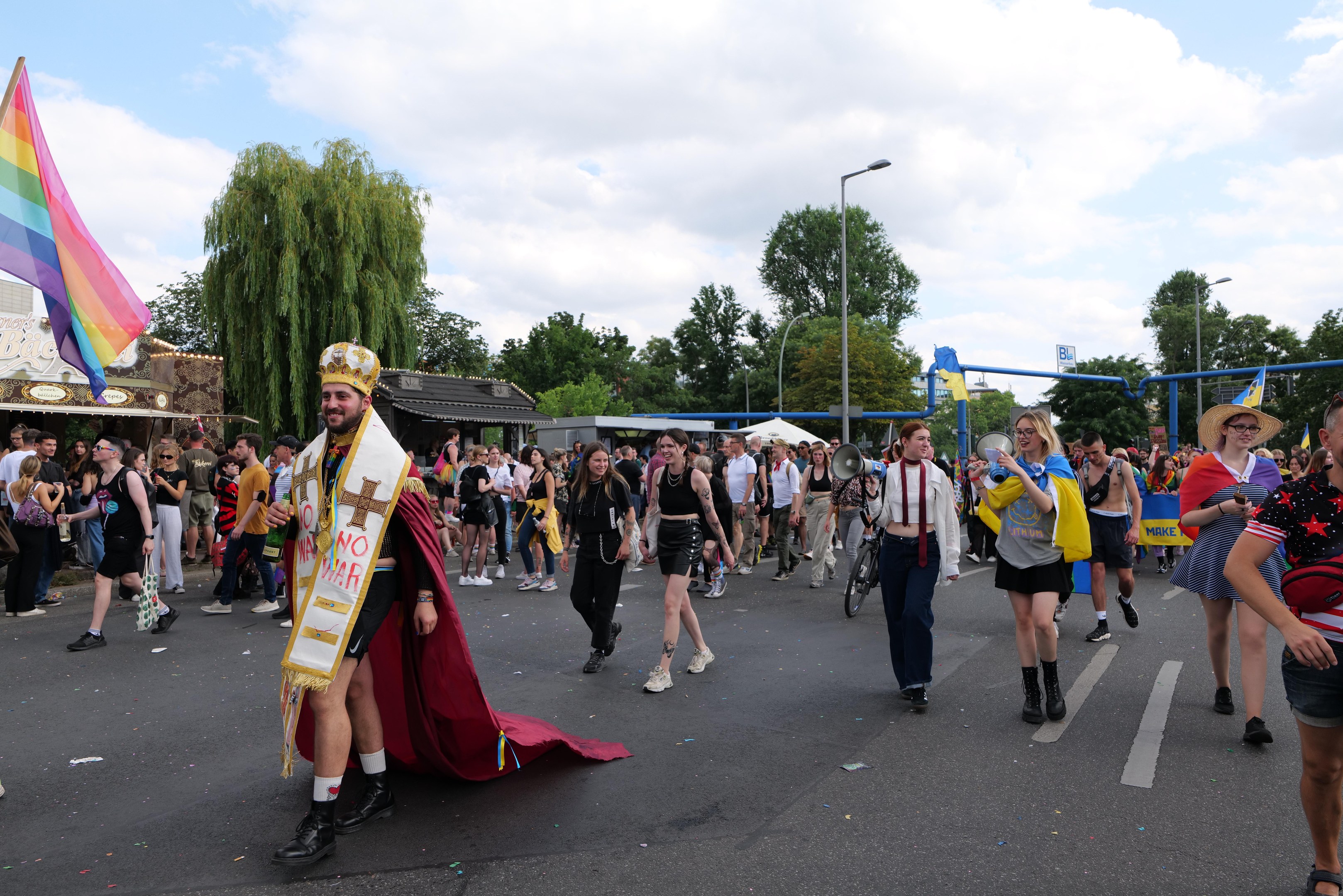 Eine Gruppe von Menschen marschiert bei der Gay Pride Parade 2018 mit einer Regenbogenflagge und Musikinstrumenten, mit Laternenmasten, Bäumen, Schuppen und einem bewölkten Himmel im Hintergrund.