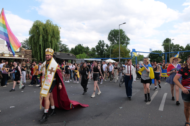 Eine Gruppe von Menschen marschiert bei der Gay Pride Parade 2018 mit einer Regenbogenflagge und Musikinstrumenten, mit Laternenmasten, Bäumen, Schuppen und einem bewölkten Himmel im Hintergrund.