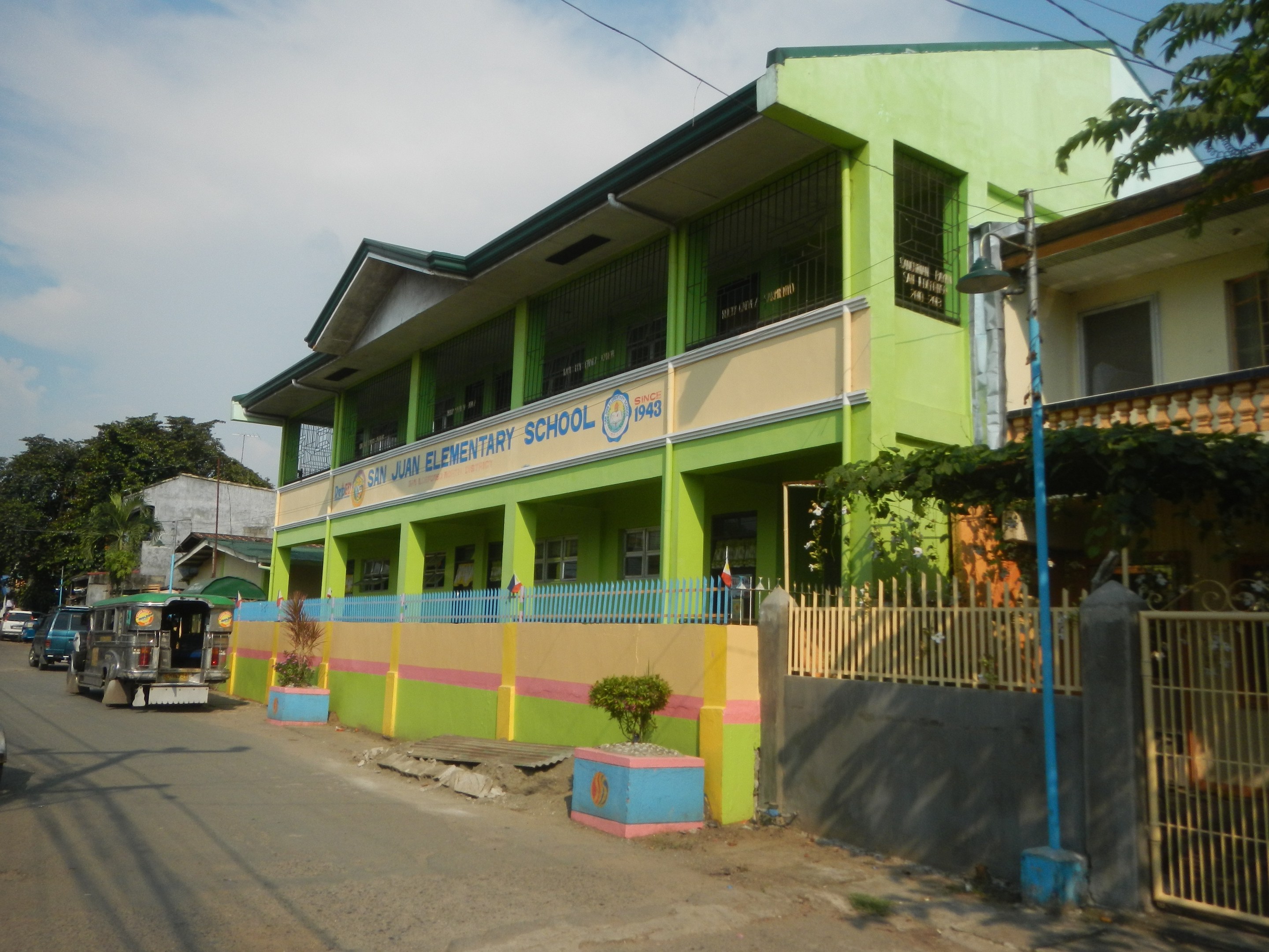 Schulgebäude mit grüner und gelber Fassade, Fahrzeuge auf der Straße, Topfpflanzen, ein Zaun, ein Tor, ein Straßenmast, Bäume, Oberleitungen und eine bewölkte Himmel.