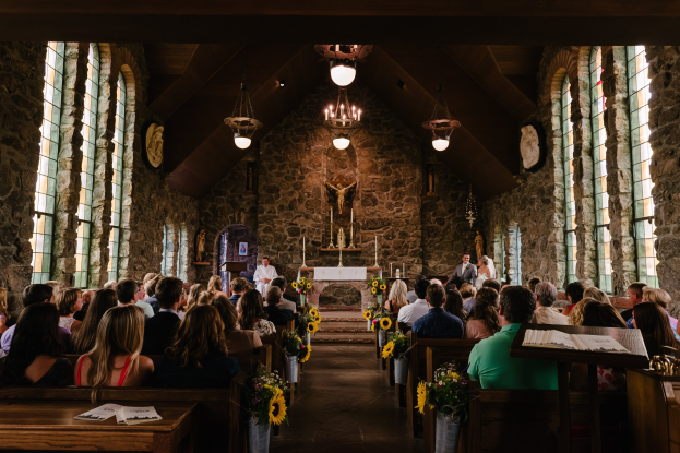 Eine Gruppe von Menschen sitzt in Kirchenbänken während einer Hochzeitszeremonie, mit Blumenschmuck, Büchern und dekorativen Gegenständen auf den Tischen, Glasfenstern an den Seiten, einer Statue und Kerzen im Hintergrund und Kronleuchtern an der Decke.