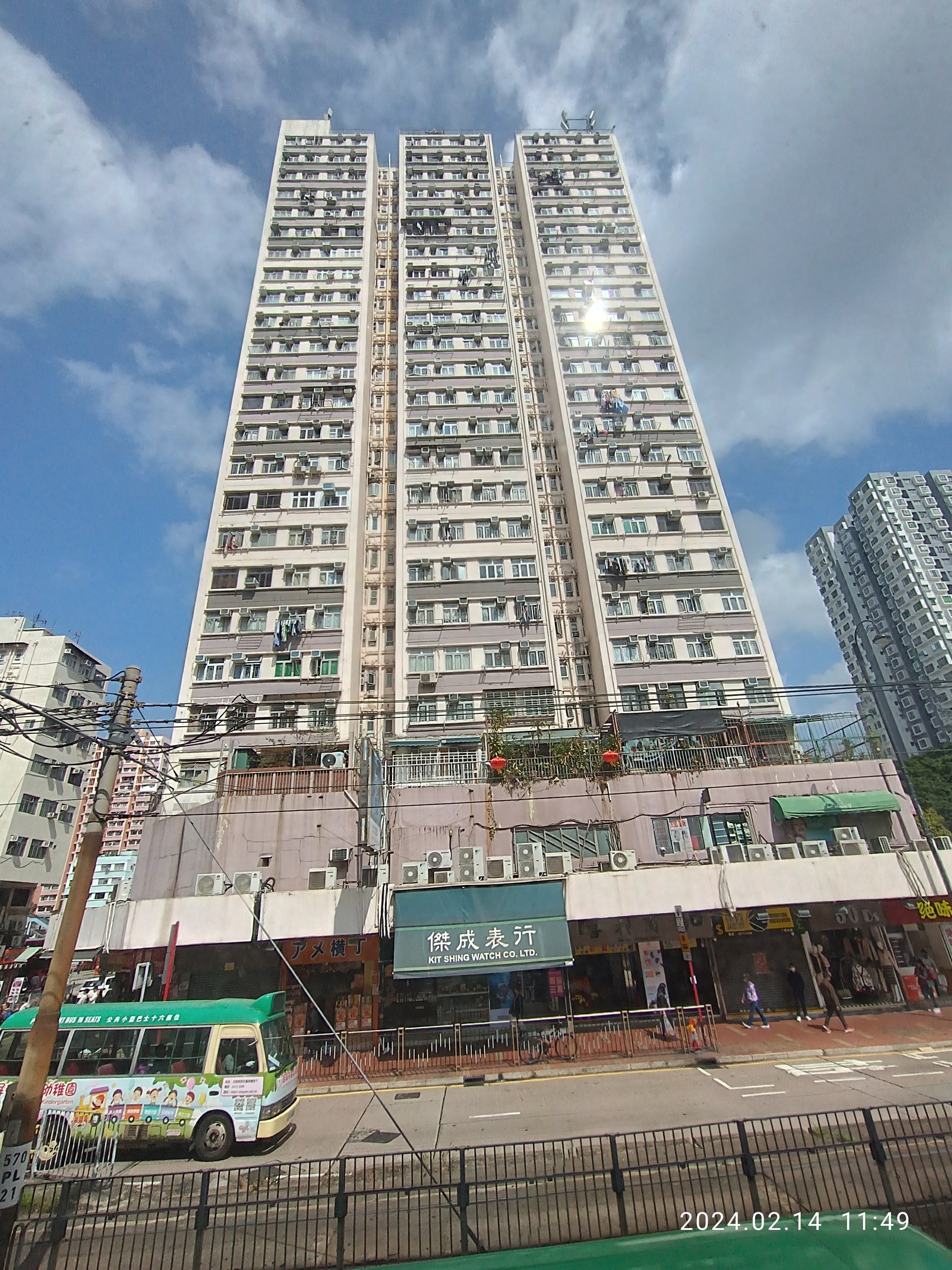 Ein hohes Wohnhaus auf einer belebten Stadtstraße umgeben von anderen Gebäuden, Fahrzeugen, Fußgängern und städtischen Elementen unter einem bewölkten Himmel.