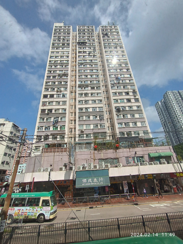 Ein hohes Wohnhaus auf einer belebten Stadtstraße umgeben von anderen Gebäuden, Fahrzeugen, Fußgängern und städtischen Elementen unter einem bewölkten Himmel.