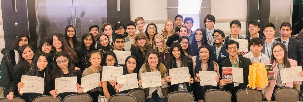 Gruppe von Menschen mit Urkunden, die zusammen in einem Raum mit Stühlen und einer Wand im Hintergrund stehen, wahrscheinlich feiern sie eine Abschlussfeier für das akademische Jahr 2019-2020.