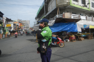 Ein Polizist in grüner und blauer Uniform mit blauer Mütze steht auf einer Straße, spricht in ein Handy, mit Fahrzeugen und Marktständen im Hintergrund und einem Gebäude im bewölkten Himmel.