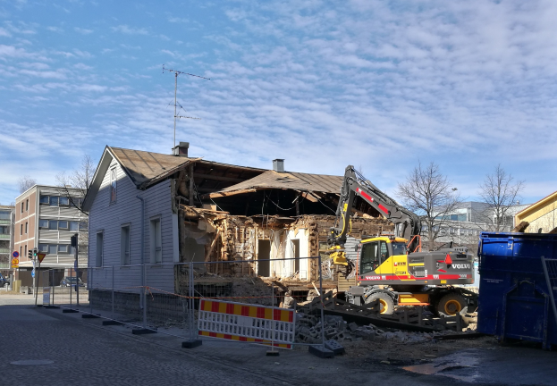 Baustelle mit einem großen Gebäude, das abgerissen wird, mit einem Kran, einem Zaun, einer Absperrung, Steinen, einem Container, Bäumen, Pfählen, einem Schild, einem Straßenpfahl, Fahrzeugen, Gebäuden mit Fenstern und einem bewölkten Himmel.