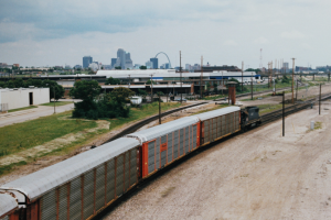 Güterzug auf Schienen neben einer Stadtlandschaft mit Gebäuden, Bäumen, Gras, Strommasten, Kabeln und einem bewölkten Himmel.