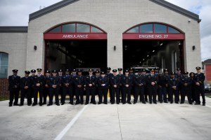 Feuerwehrleute in Uniform und Mützen stehen vor einer Feuerwache mit Gras, geparkten Fahrzeugen, Gebäuden und einem klaren blauen Himmel im Hintergrund.