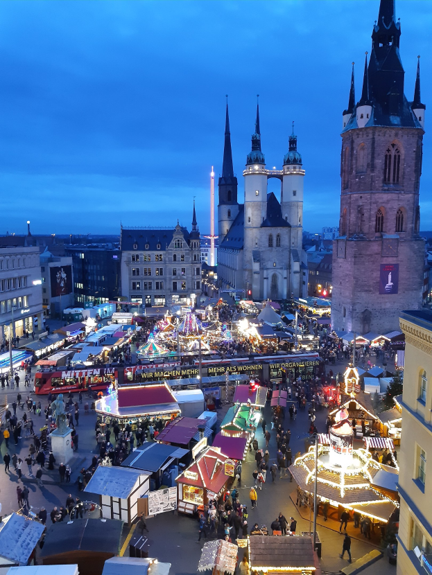 Ein belebter Weihnachtsmarkt in Köln, Deutschland bei Nacht mit Menschen, die von Stand- und Gebäudelichtern beleuchtet werden, unter einem bewölkten Himmel.