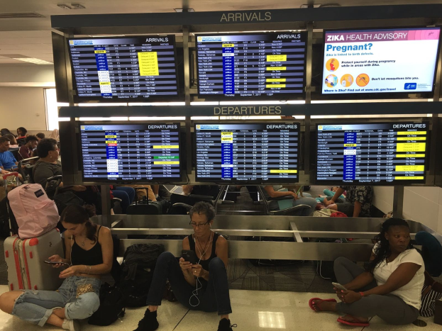 Menschen sitzen auf einem Flughafenboden, einige halten Handys, mit verstreuten Taschen und Bildschirmen, die Zika-Virus-Informationen anzeigen, im Hintergrund.