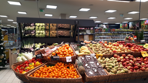 Ein Ladeneinrichtungsbild mit Körbüssern, Obst und Gemüsen in Körbüssern mit Preisschildern, einem dekorierten Weihnachtsbaum links und Regalen mit anderen Artikeln im Hintergrund.