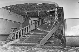 Ein Schwarz-Weiß-Foto einer Treppe mit einem Geländer, umgeben von einer Wand mit einer Tafel und ein paar verstreuten Gegenständen am Boden.