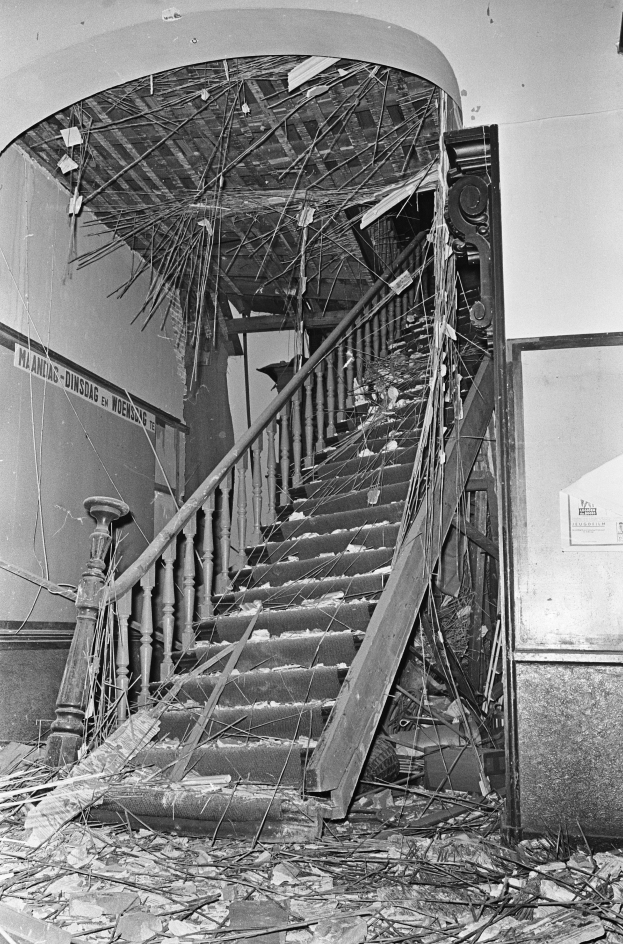 Ein Schwarz-Weiß-Foto einer Treppe mit einem Geländer, umgeben von einer Wand mit einer Tafel und ein paar verstreuten Gegenständen am Boden.