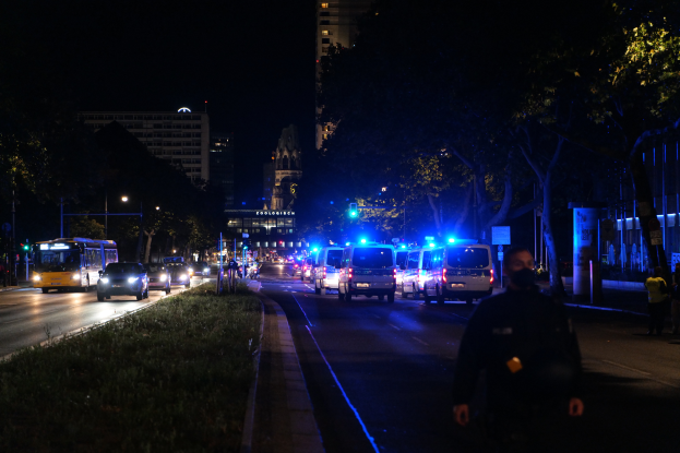 Eine nächtliche Straßen Szenerie mit Polizeiwagen und Bussen, Menschen auf dem Gehweg, Gras auf der linken Seite, Bäume und Gebäude im Hintergrund und Laternen, die die Straße beleuchten.