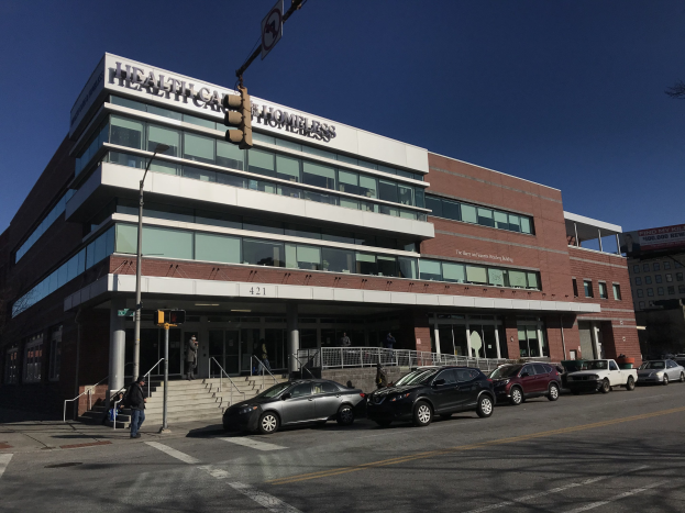 Großes Gebäude mit der Aufschrift "New York City Health Care Homeless Center" und parkenden Autos, Fußgängern, Straßenmöblierung und Bäumen unter einem klaren blauen Himmel.