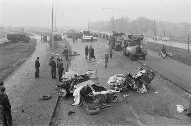 Schwarz-weiß-Szene eines Autounfalls am Straßenrand mit mehreren Fahrzeugen, einer Gruppe von Menschen in der Nähe des beschädigten Autos, Laternenpfähle, Bäume und den Himmel im Hintergrund.