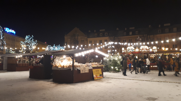 Lebendiger Weihnachtsmarkt an einem verschneiten Abend mit Menschen, Ständen, Pflanzen, Bäumen, Gebäuden und Schildern unter einem bewölkten Himmel.