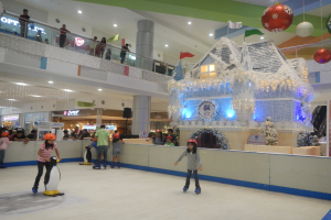 Eine Gruppe von Menschen, die auf einer Eisbahn in einem Einkaufszentrum Schlittschuh laufen, umgeben von einem hausähnlichen Aufbau, der mit Lichtern und festlichen Dekorationen geschmückt ist, mit Zuschauern und zusätzlichen Dekorationen, die eine festliche Atmosphäre schaffen.