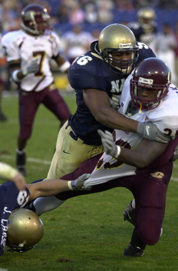 Zwei Fötballspieler in Helmen und Uniformen kollidieren auf dem Feld, während andere zuschauen.