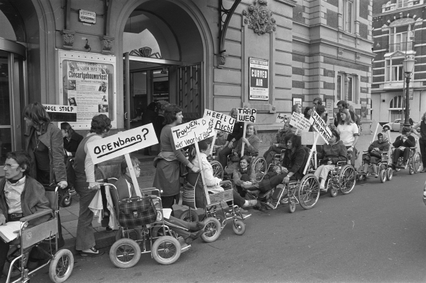 Eine Gruppe von Menschen im Rollstuhl mit Schildern auf der Straße, mit Gebäuden im Hintergrund und einem Laternenmast auf der rechten Seite, in Schwarz-Weiß.