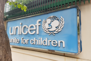 Das UNICEF-Logo ist auf der Wand eines Gebäudes zu sehen, mit einem Baumstamm links daneben und einer Tafel an der Wand, vor einem Hintergrund aus Bäumen und einem klaren blauen Himmel.