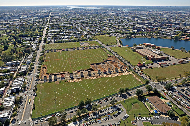 Luftaufnahme einer Stadt mit dichtem Baumbestand, Grünflächen, Straßen mit Fahrzeugen, einem Gewässer und einem bewölkten Himmel, die einen zentralen, leeren Platz für einen Park zeigt.