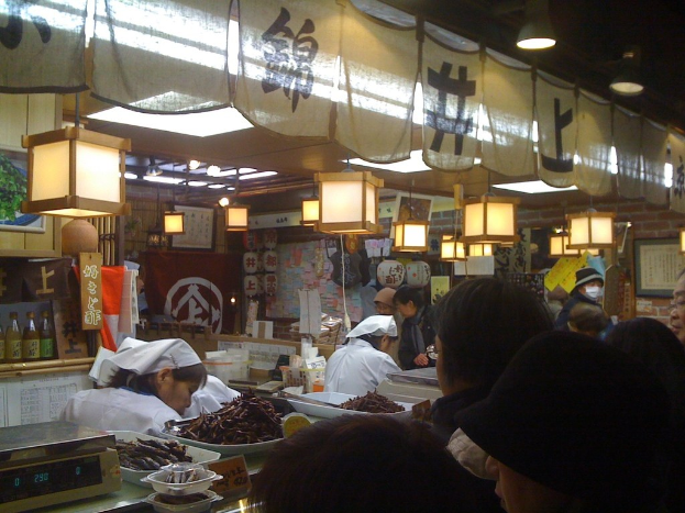 Restaurantmitarbeiter bereiten Bestellungen hinter einer Theke vor, während Kunden davor stehen und Flaschen mit Soßen, Menükarten und hängende Kleidungsstücke im Hintergrund zu sehen sind.