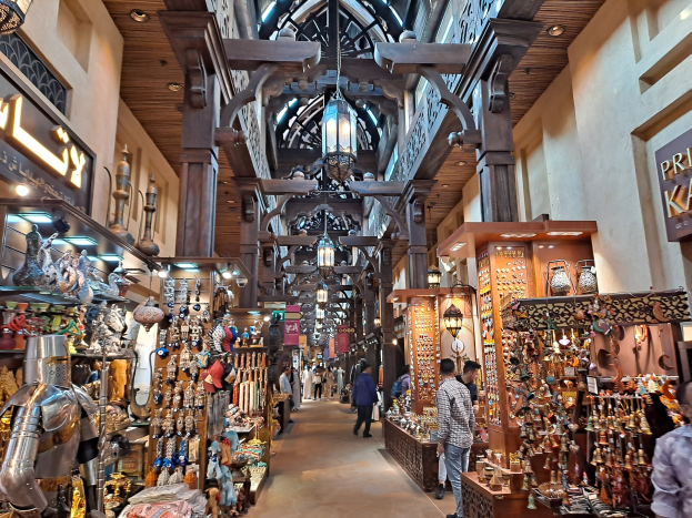 Ein belebter Dubai-Basar mit Menschen, die zwischen Regalen voller Waren hindurchgehen, beleuchtet von Deckenlampen und mit Texttafeln an den Wänden.