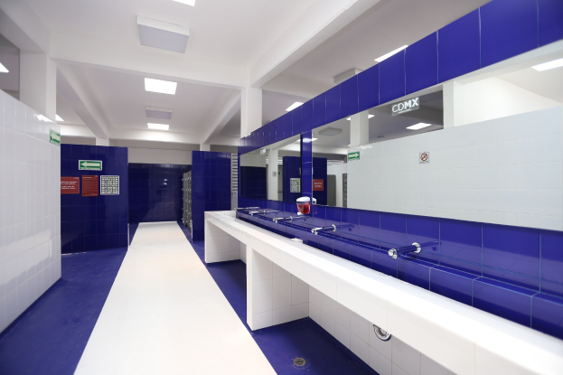 Öffentliche Toilette mit blauen Fliesenwänden, weißen Arbeitsplatten, Waschbecken mit Wasserhähnen, einer Plattform mit einer Flasche und anderen Gegenständen, Informationsschildern an den Wänden, Deckenleuchten und Stützpfeilern.