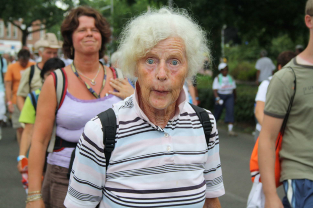 Eine ölte Frau mit einem Überraschten Gesichtsausdruck geht die Straße entlang, umgeben von Menschen, einige tragen Taschen, mit Bäumen, Gebäuden und einem klaren Himmel im Hintergrund.