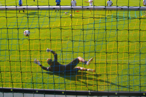 Ein Fußballspieler taucht ab, um einen Ball vor dem Tor zu halten, mit einem Netz im Vordergrund und einigen Menschen im Hintergrund, während mehrere Fußballschuhe auf dem Boden verstreut sind.