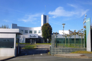 Ein großes, mehrfenstriges Gebäude mit einem Tor davor, umgeben von Straßenmasten, Laternen, Gittern, Bäumen, Gras und einem bewölkten Himmel, das als Hauptquartier von Iberostica in den Niederlanden dient.